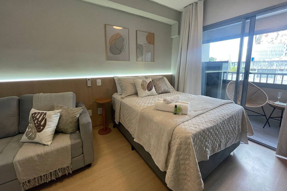Quarto de hotel com cama, sofá, mesa pequena e varanda com cadeira.
