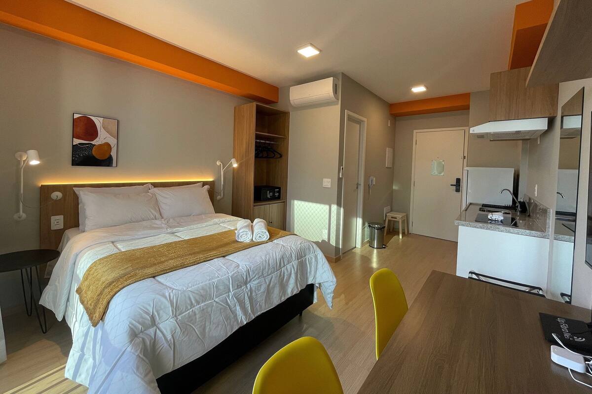 Quarto de hotel com cama, mesa de cabeceira, luminária e uma pequena cozinha.