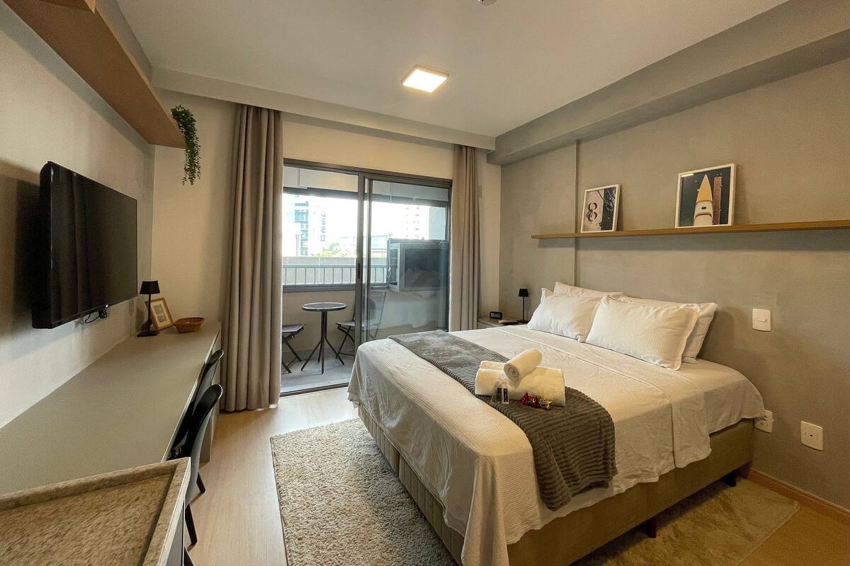 Quarto de hotel com cama, escrivaninha, televisão, varanda com vista e uma pequena mesa.