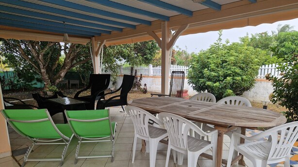 An outdoor patio with a wooden table and chairs, surrounded by greenery and a white fence. - House with 3 air-conditioned bedrooms. (Lamentin)