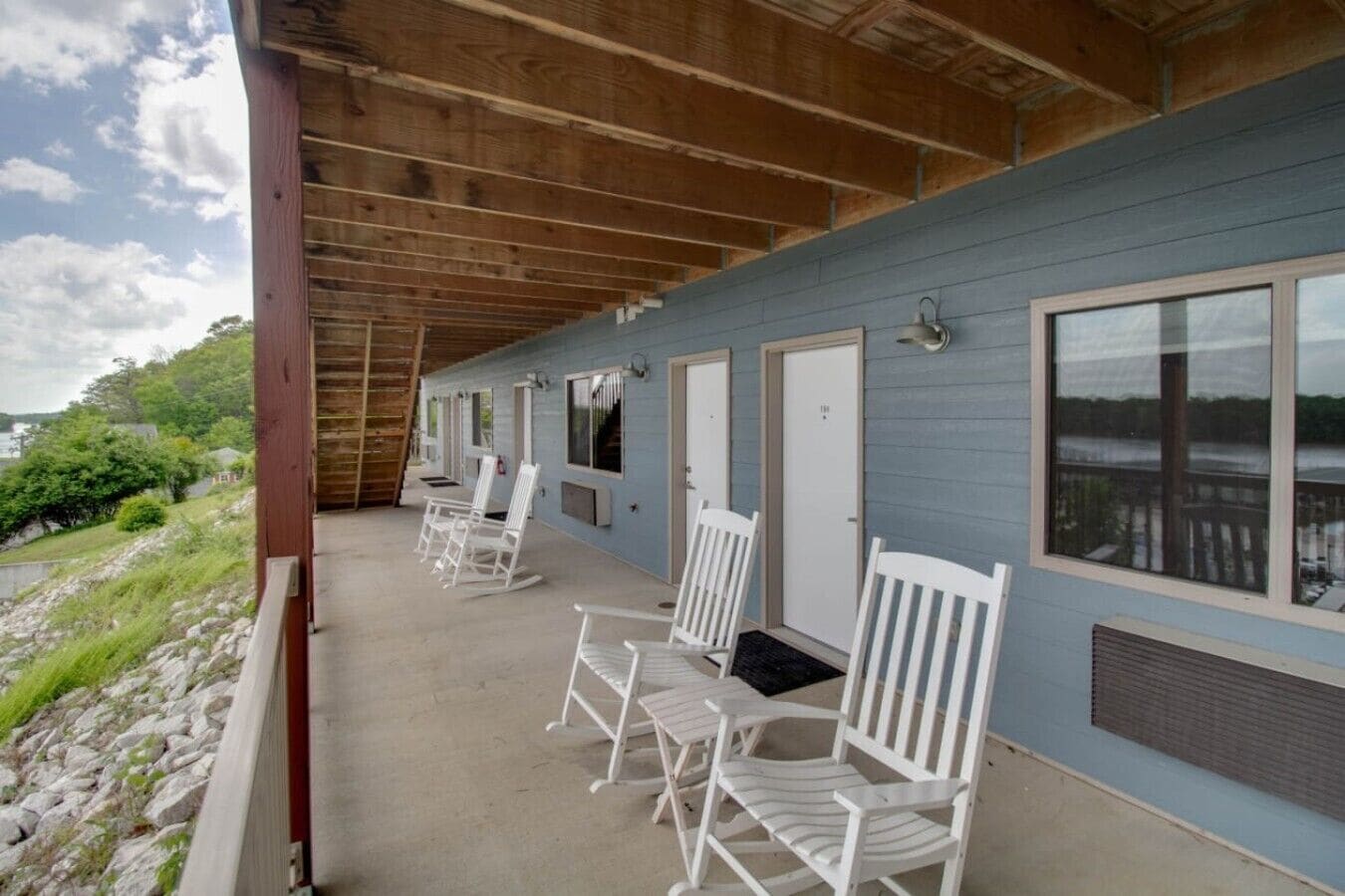 A covered porch with white rocking chairs, a wooden ceiling, and a view of a lake.