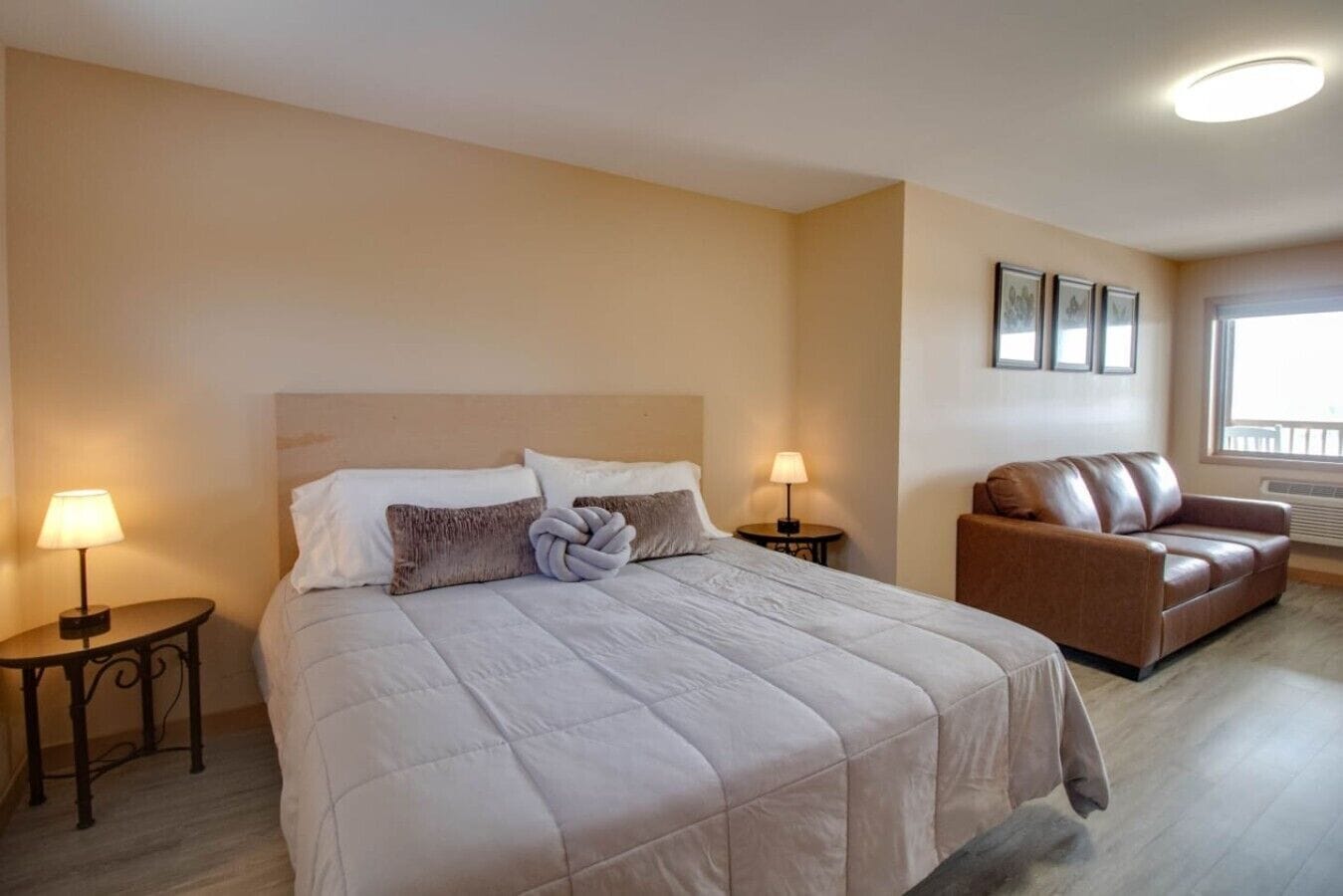Une chambre d’hôtel avec un lit, deux tables de chevet avec des lampes, un canapé en cuir marron et une fenêtre avec des stores.