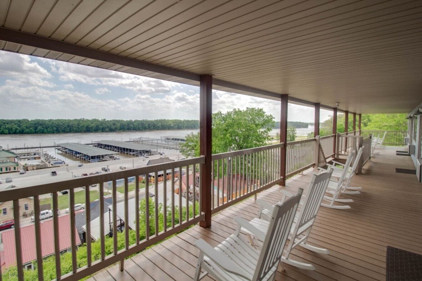 A covered balcony with wooden flooring and white rocking chairs, overlooking a parking lot and a body of water.