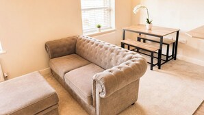 A living room with a beige sectional sofa, a small wooden table, and a potted plant. - Modern Apartment in the Heart of Bourne | Sleeps 4 (Bourne)