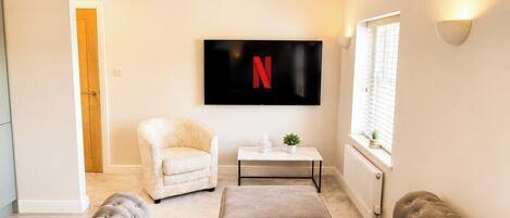 En stue med sofa, lænestol, skammel, et fladskærms-tv og et lille bord med en plante.