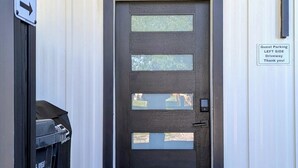 Une porte dotée d’un cadre noir et de panneaux en verre dépoli, un panneau indiquant les places de stationnement réservées aux visiteurs sur le mur, et un paillasson avec l’inscription « Bonjour » à l’entrée.
