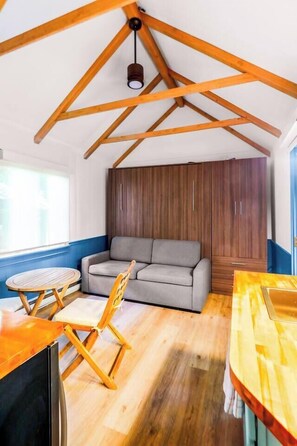 A room with a wooden ceiling, a grey sofa, a wooden dining table, and a wooden chair. - Charming Private Cottage w/ Deck, Murphy Bed (Provincetown)