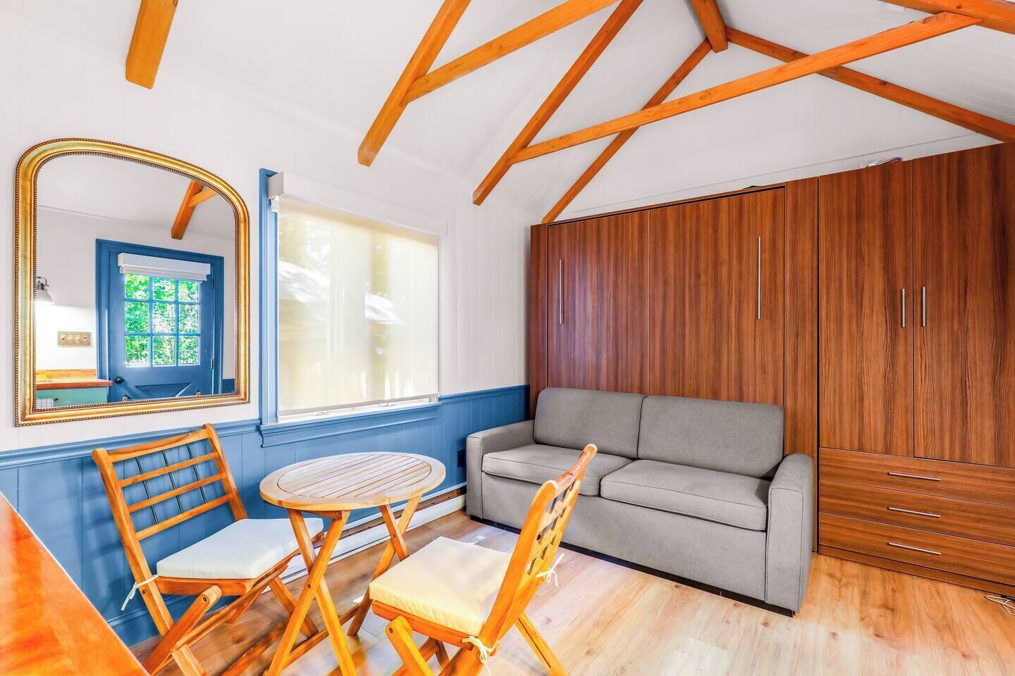 A room with a sofa, two wooden chairs, a small round table, a mirror, and a wardrobe.