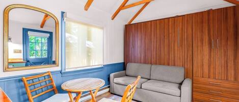 Una habitación con un sofá, dos sillas de madera, una mesa redonda pequeña, un espejo y un armario.