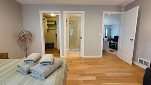Ein Schlafzimmer mit einem Bett, zwei Handtüchern, einem Standventilator sowie ein Badezimmer mit Dusche und Toilette, das durch eine offene Tür sichtbar ist.