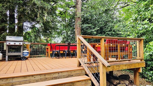 Eine Holzterrasse mit Grill, roter Sitzgelegenheit und einem eingezäunten Bereich mit Blick auf Bäume.