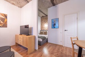 Ein modernes Zimmer mit einem Holzbüro, einem Flachbildfernseher, einem Bett vor einer Ziegelwand, einem Esstisch mit Stuhl und einer Tür.