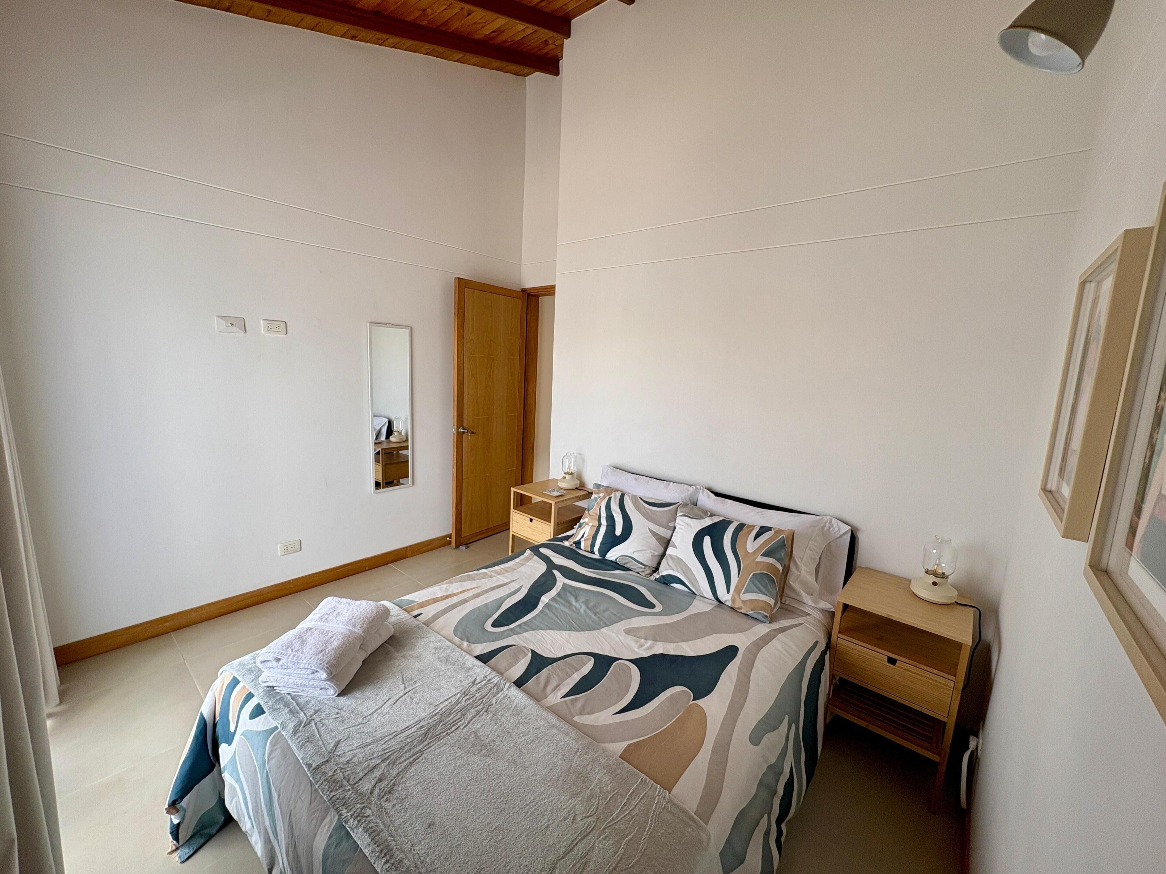 A bedroom with a bed, bedside table, nightstand, mirror, wardrobe, and a framed picture on the wall.