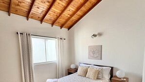 A bedroom with a wooden ceiling, a bed with patterned bedding, a nightstand with a lamp, and a window with curtains.