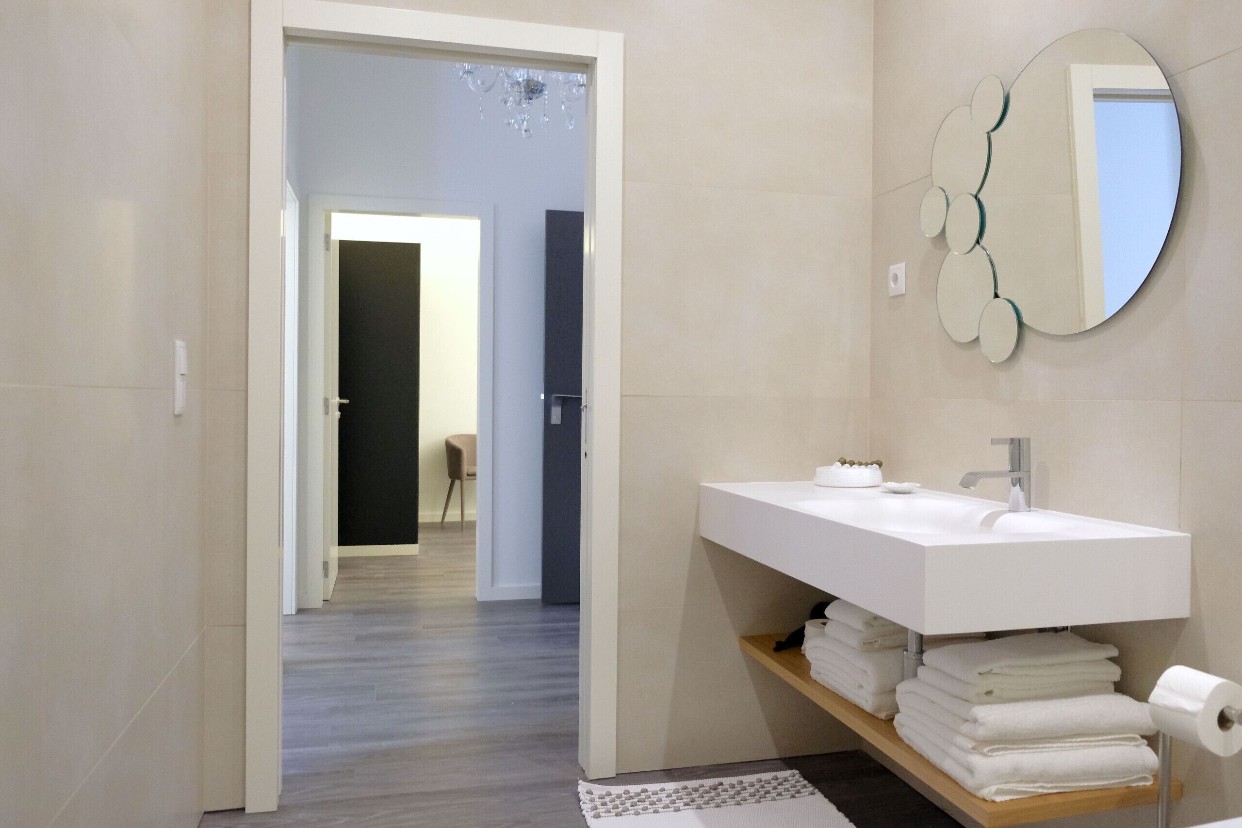 A modern bathroom with a white sink, a mirror with a circular frame, and a wooden shelf with towels.