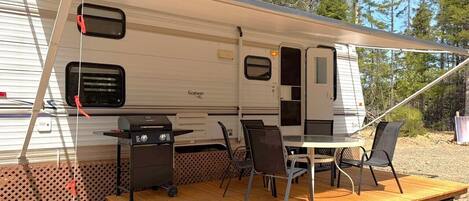 A recreational vehicle with a covered patio area, a grill, and a table with chairs.
