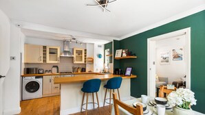 Une cuisine compacte avec des meubles en bois, un lave-linge, deux tabourets de bar et un coin repas avec une table dressée pour deux personnes.