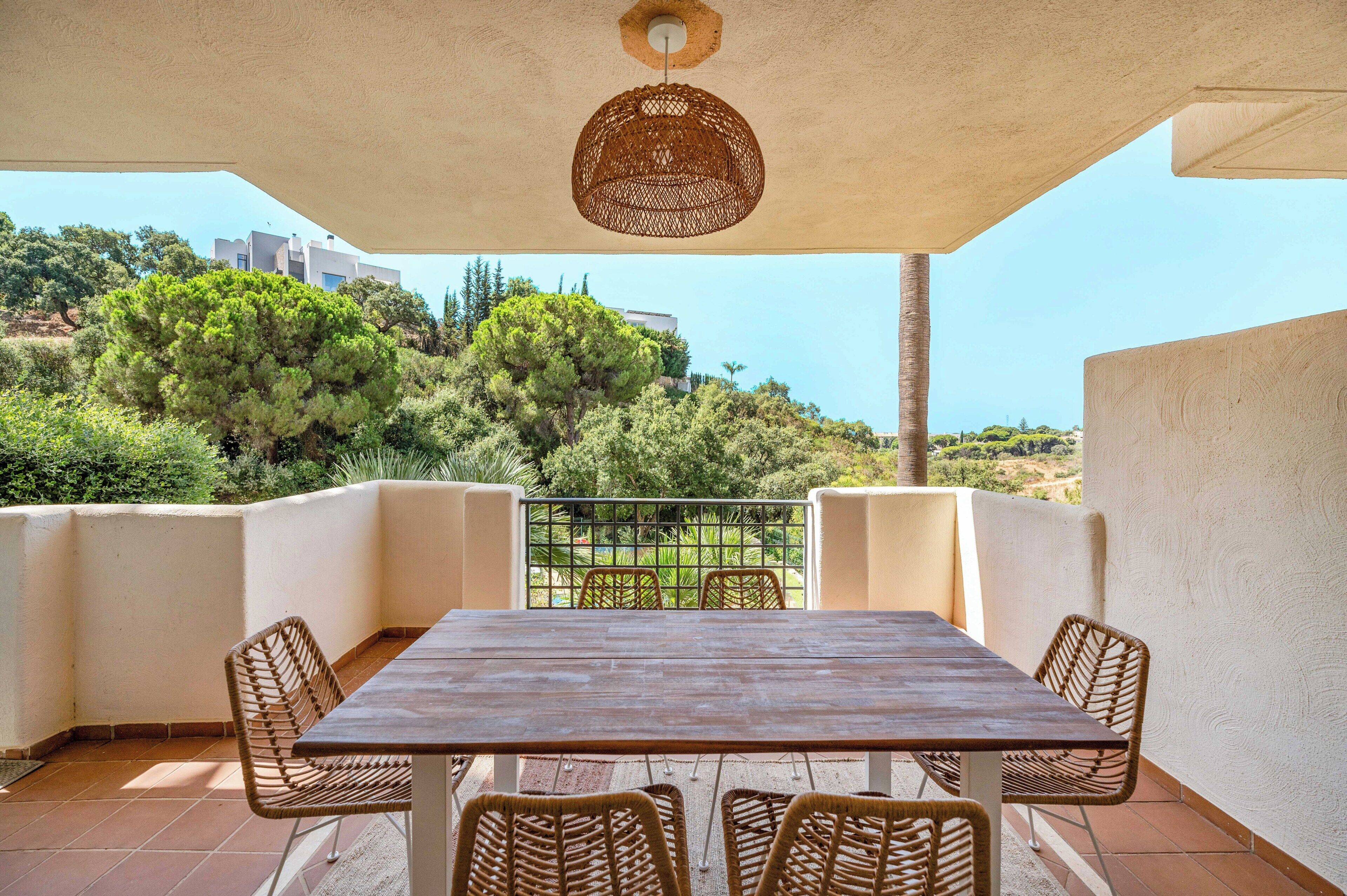 Une terrasse extérieure couverte, avec une table et des chaises, un luminaire suspendu et une vue sur la verdure et les bâtiments.