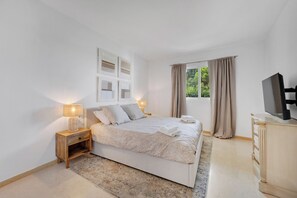 A bedroom with a bed, bedside lamps, a TV, and a window with curtains. - LXR Elviria Hills 2-Bedroom Apartment with Heated Pool (Marbella)