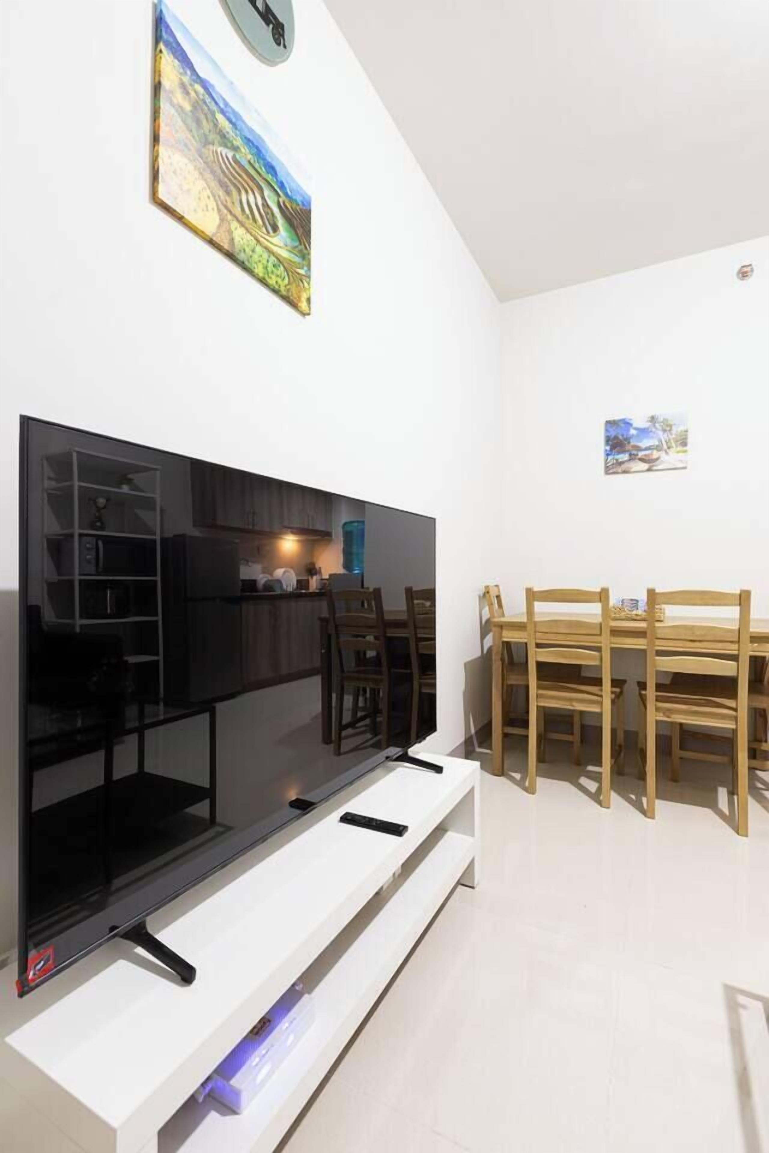 A modern living room with a flat-screen TV, wooden dining table, and chairs, with a kitchen area in the background.