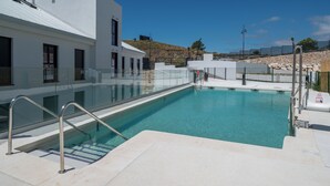 Una piscina con barrera de vidrio, un edificio blanco y un muro de piedra al fondo.