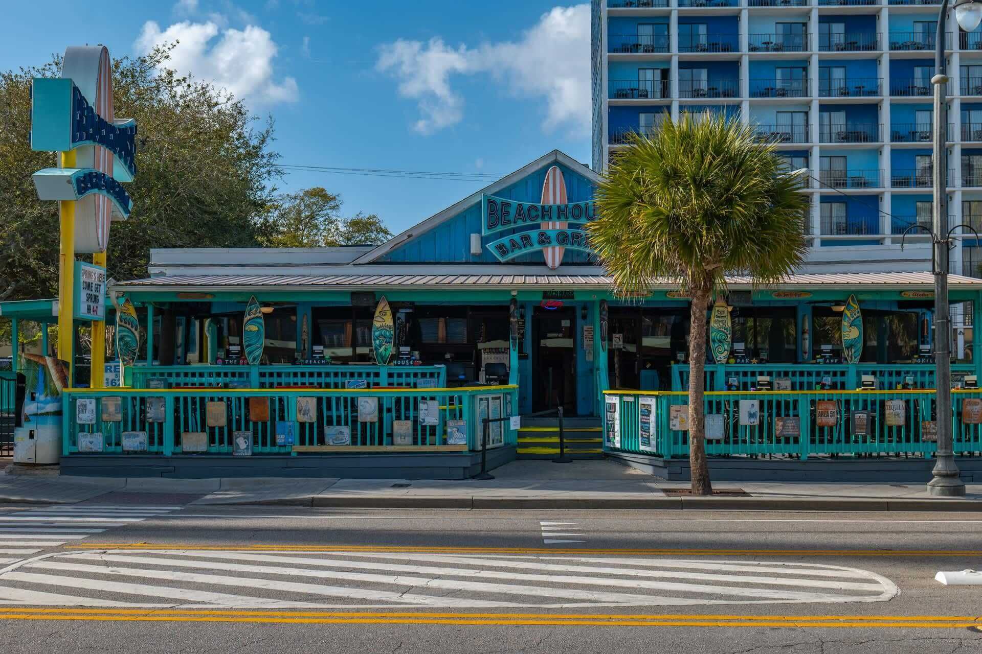 A beachfront bar and grill with a blue facade, a palm tree in front, and a building with balconies in the background.