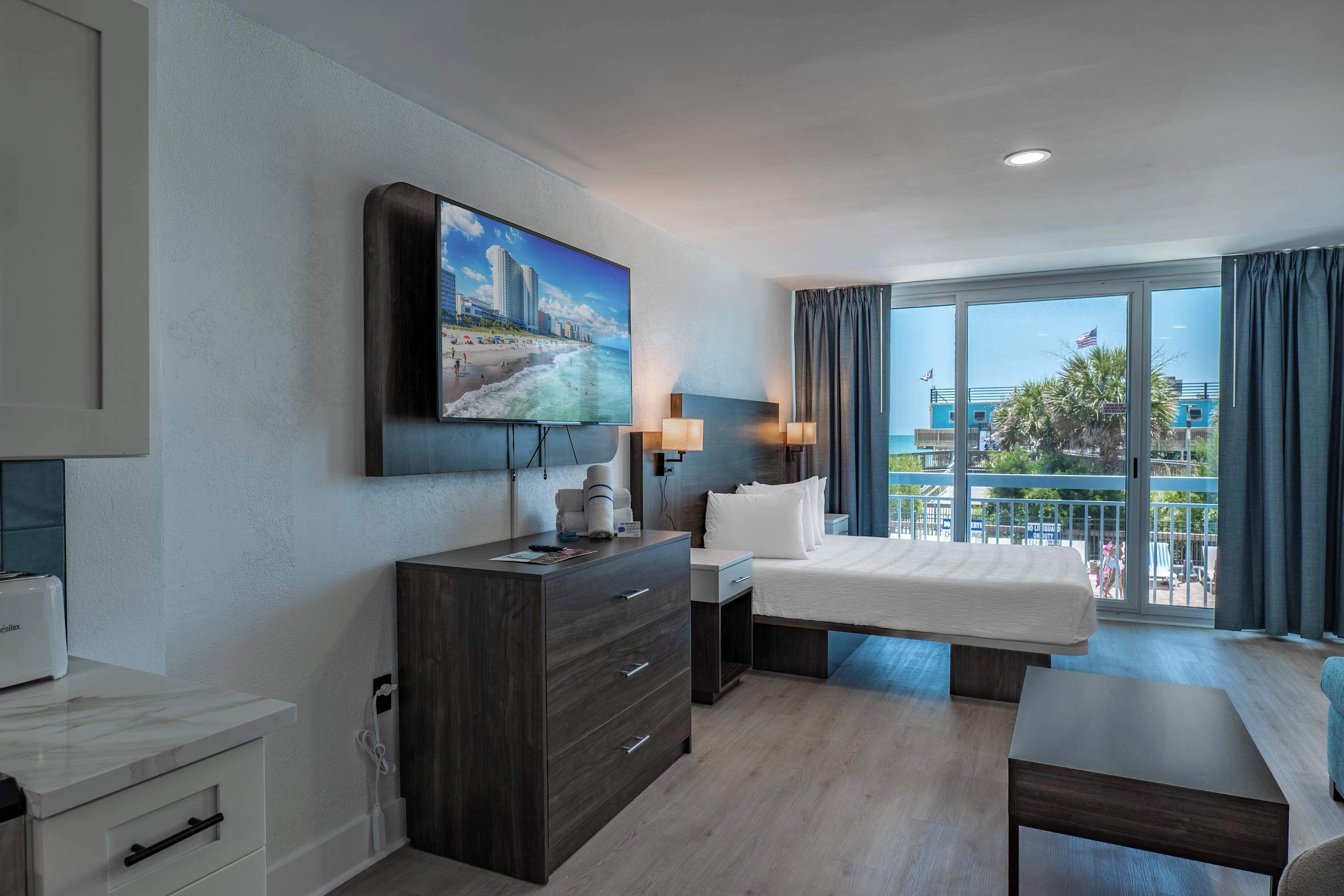 A hotel room with a bed, a desk, a TV, and a balcony with a view of buildings and palm trees.