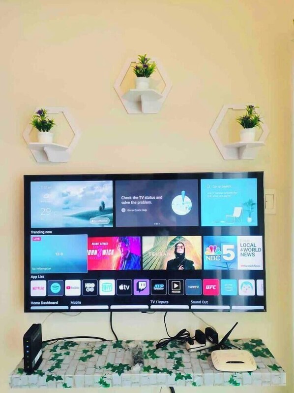 A flat-screen TV mounted on a wall with a home dashboard interface, surrounded by hexagonal shelves with potted plants.