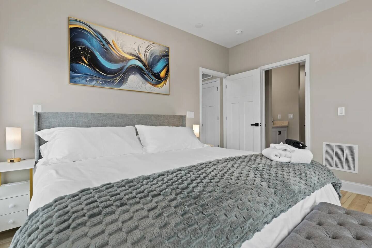 A bedroom with a bed, a grey blanket, a grey headboard, a bedside table with a lamp, a wall-mounted abstract painting, and a door leading to another room.