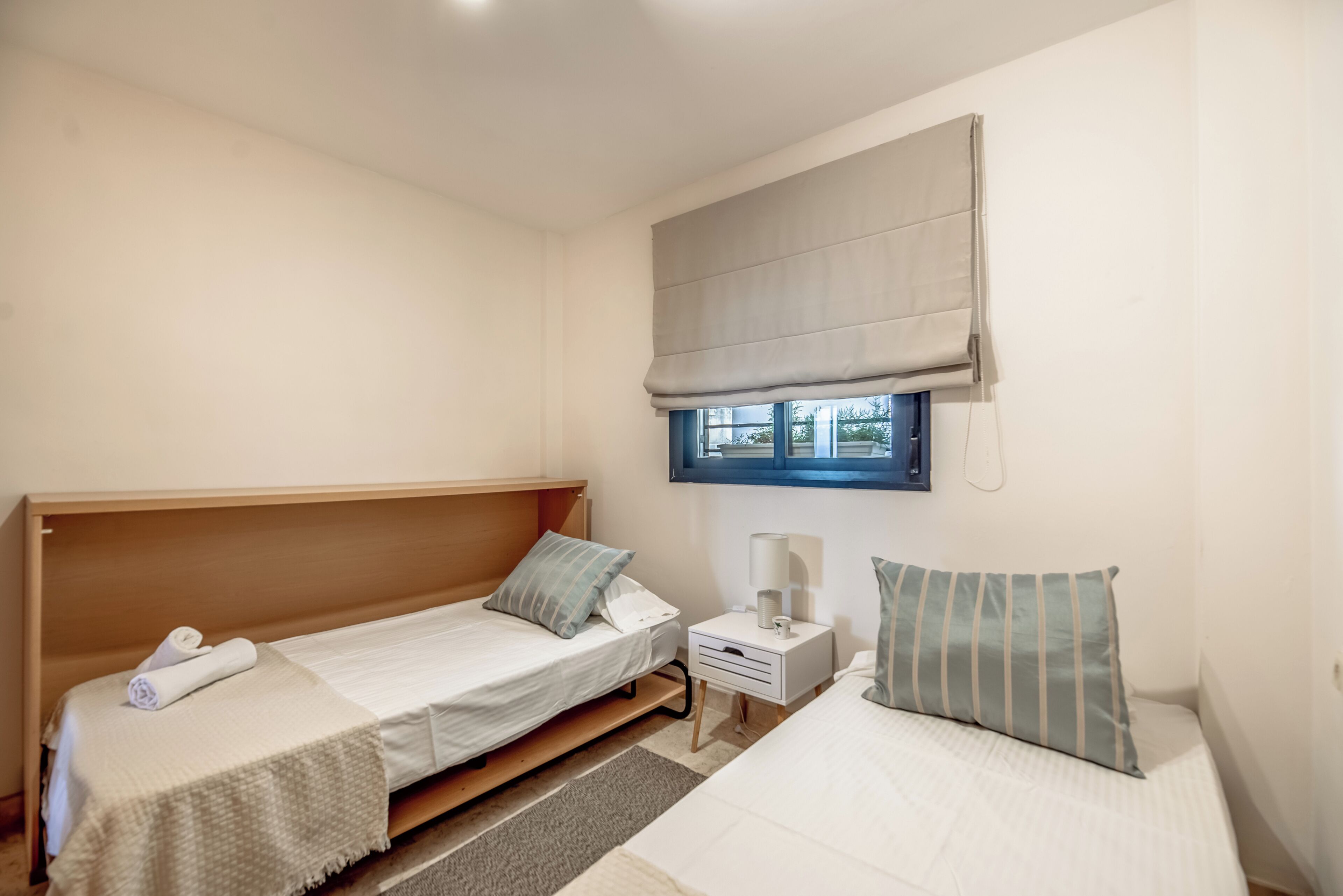 A room with two single beds, a nightstand with a lamp, a window with a blind, and a wooden headboard.