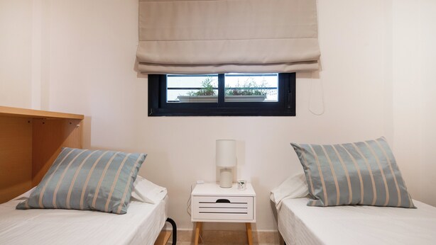 A small room with two beds, a nightstand with a lamp, and a window with a view of greenery outside. - LXR Hollywood Hills 2-Bedroom: Stunning Views (Mijas)