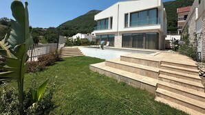 Uma casa moderna de dois andares com piscina, rodeada por um jardim bem cuidado e uma paisagem acidentada.