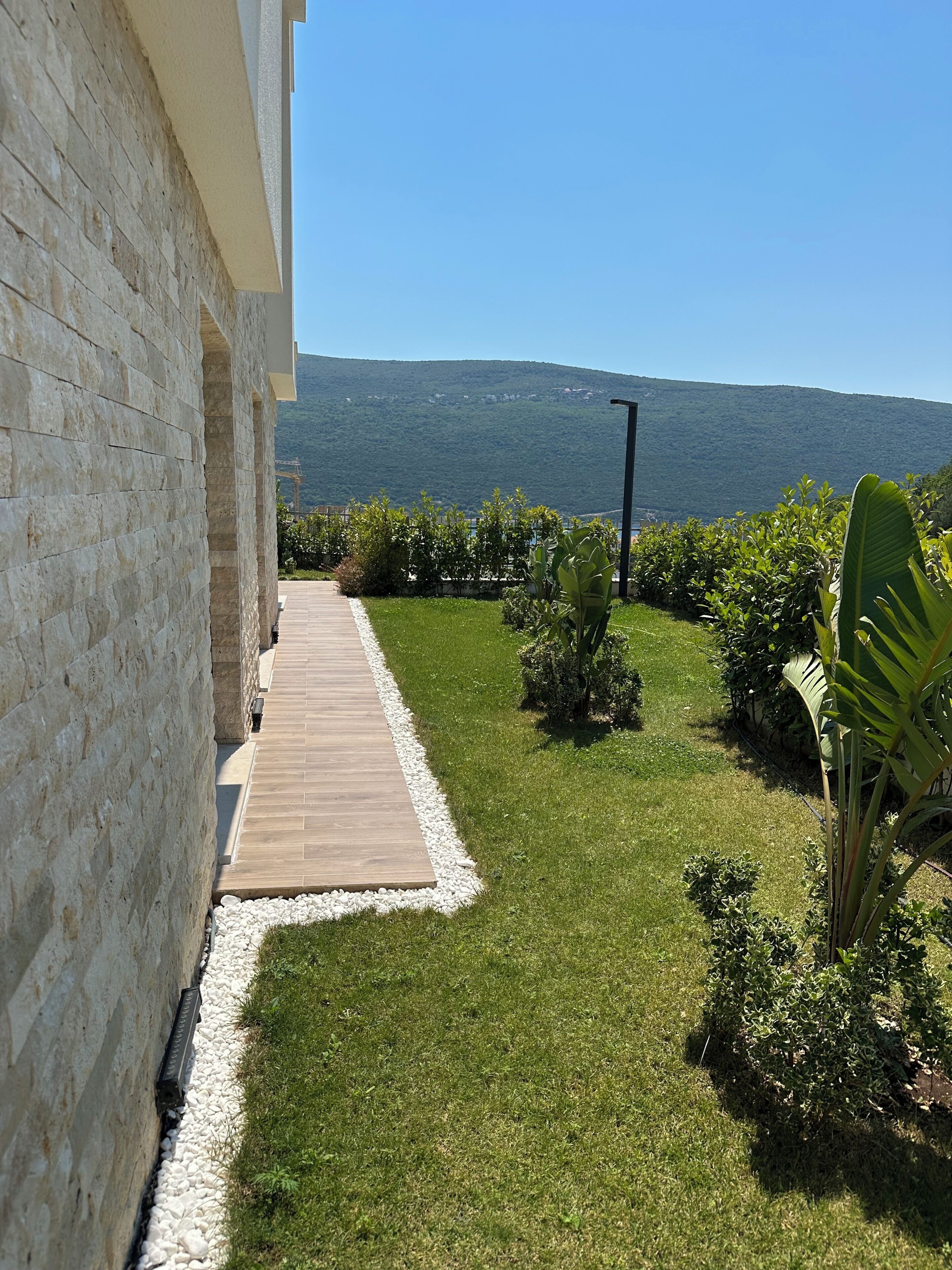 A paved pathway leading to a building with a stone facade, surrounded by a well-maintained grassy area with various plants and a view of a hill in the distance.