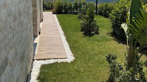 Um caminho pavimentado conduz a um edifício com fachada de pedra, rodeado por uma área ajardinada bem cuidada com diversas plantas e vista para uma colina ao longe.