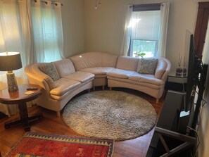 Ein Wohnzimmer mit einem Ecksofa, einem runden Teppich, einem kleinen Tisch mit Lampe und einem Fernseher.