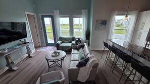 A living room with a TV, sofa, coffee table, and a dining area with a kitchen counter and chairs. - Tranquility by the Beach - Modern West End Escape - Ocean View (Galveston)