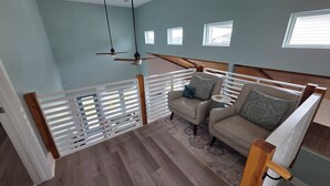 Una habitación con ventilador de techo, un sofá con cojines estampados, una mesita redonda y vistas a edificios a través de las ventanas.