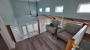 Ein Zimmer mit Deckenventilator, einer Couch mit gemusterten Kissen, einem kleinen runden Tisch und Blick auf Gebäude durch die Fenster.