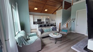A modern living room with a sofa, armchair, and coffee table, a kitchen in the background, and a staircase. - Tranquility by the Beach - Modern West End Escape - Ocean View (Galveston)