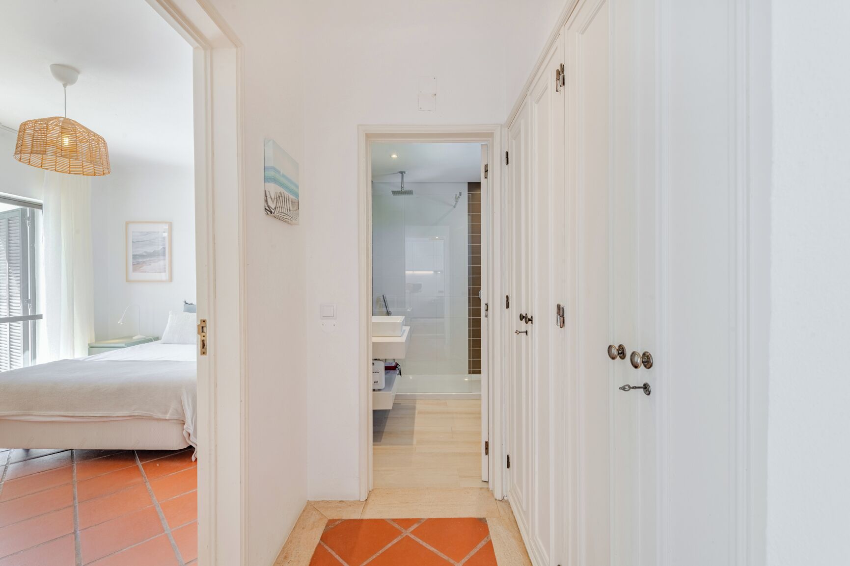 Corredor que dá para um quarto com uma cama, luminária pendente e uma porta com olho de fechadura.