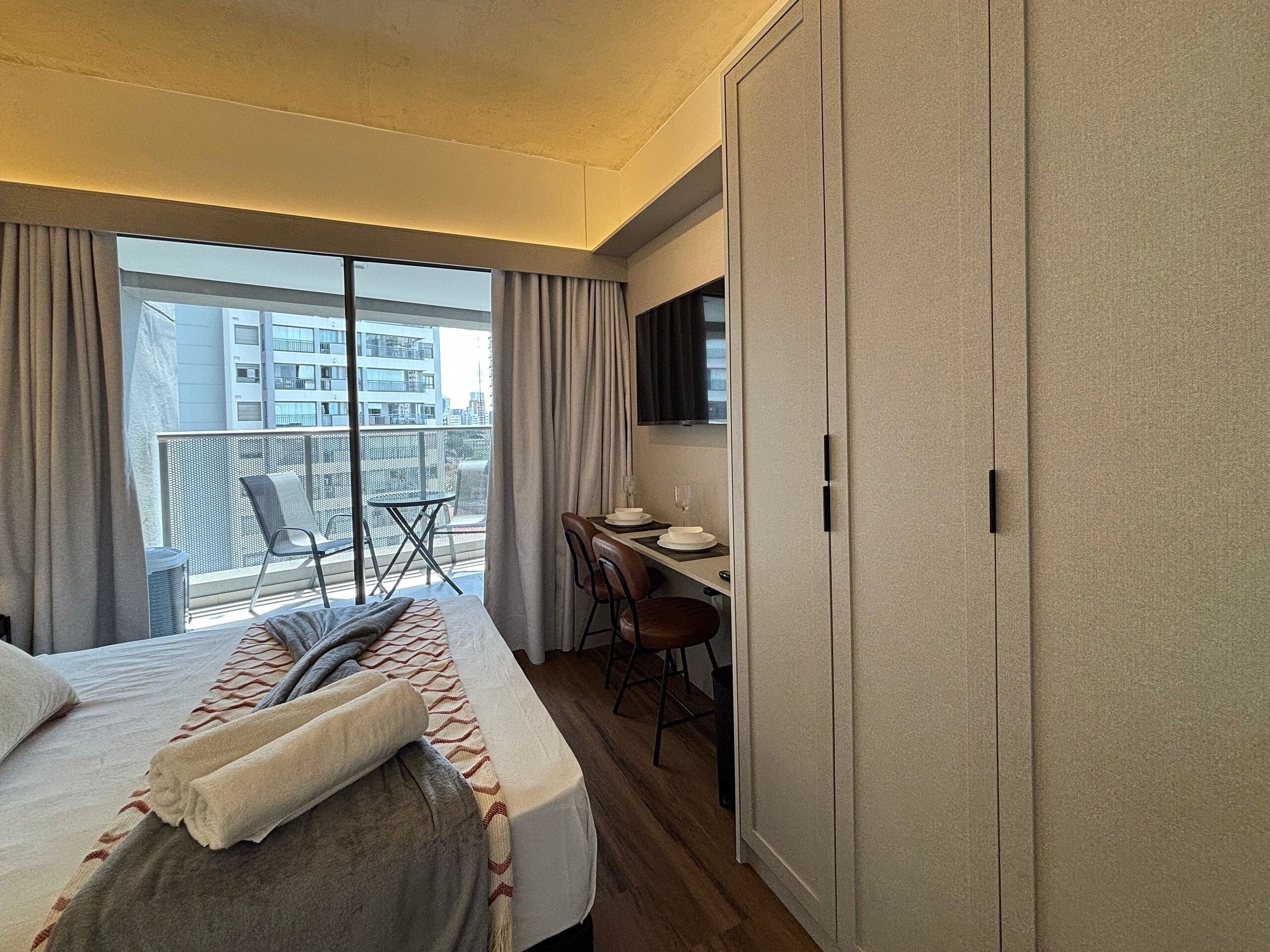A hotel room with a bed, a desk, a chair, a wardrobe, and a balcony with a view of buildings.