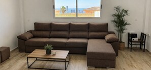 A living room with a sectional sofa, a coffee table, a potted plant, and a view of the cityscape through a window.