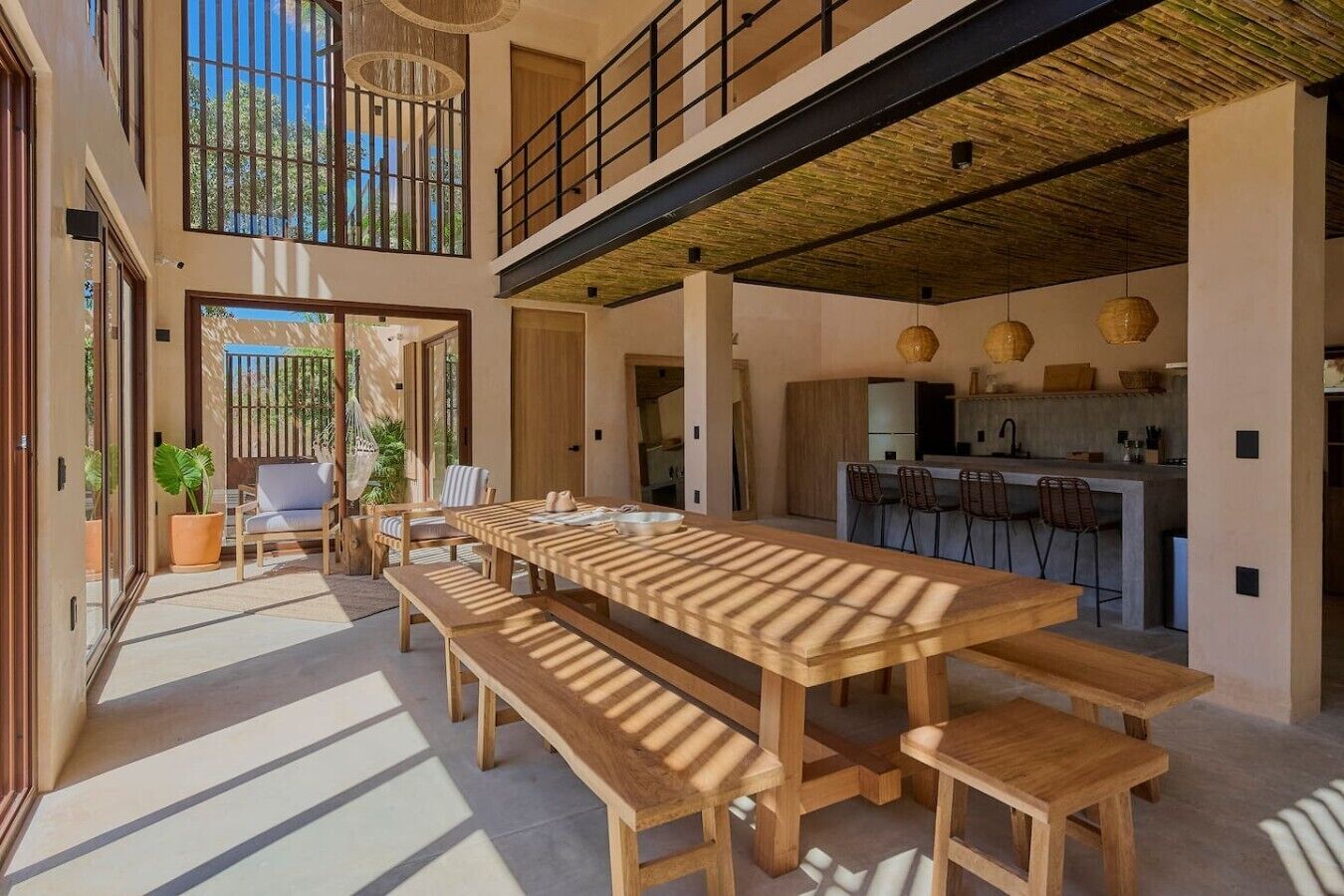 A spacious indoor area with a long wooden table and benches, a kitchen area, and a view through a large window.