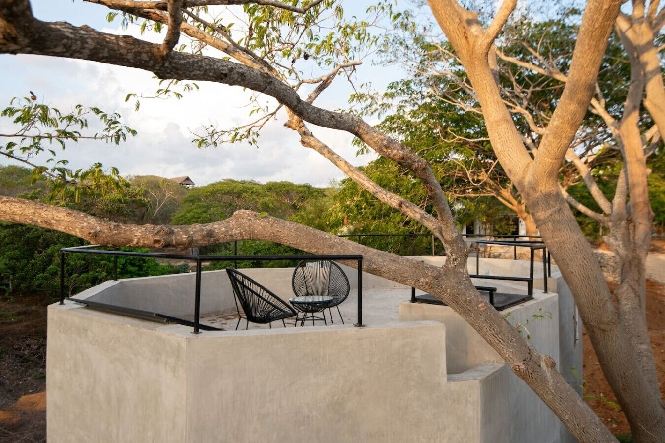 A concrete outdoor seating area with metal chairs and a table, surrounded by trees and greenery.