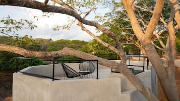 Un espacio de descanso al aire libre con piso de concreto, sillas y una mesa de metal, rodeado de árboles y follaje.