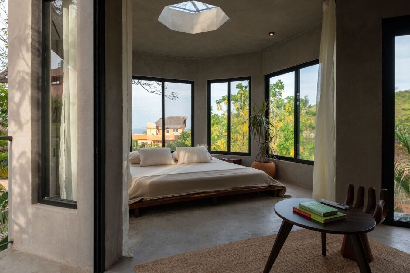 Ein Schlafzimmer mit einem großen Bett, einem runden Tisch mit Büchern und Blick auf Grünflächen und Gebäude.