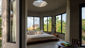 A bedroom with a large bed, a round table with books, and a view of greenery and buildings outside. - 2 albercas - Vista al Mar - Brutal Verde (Brisas de Zicatela)