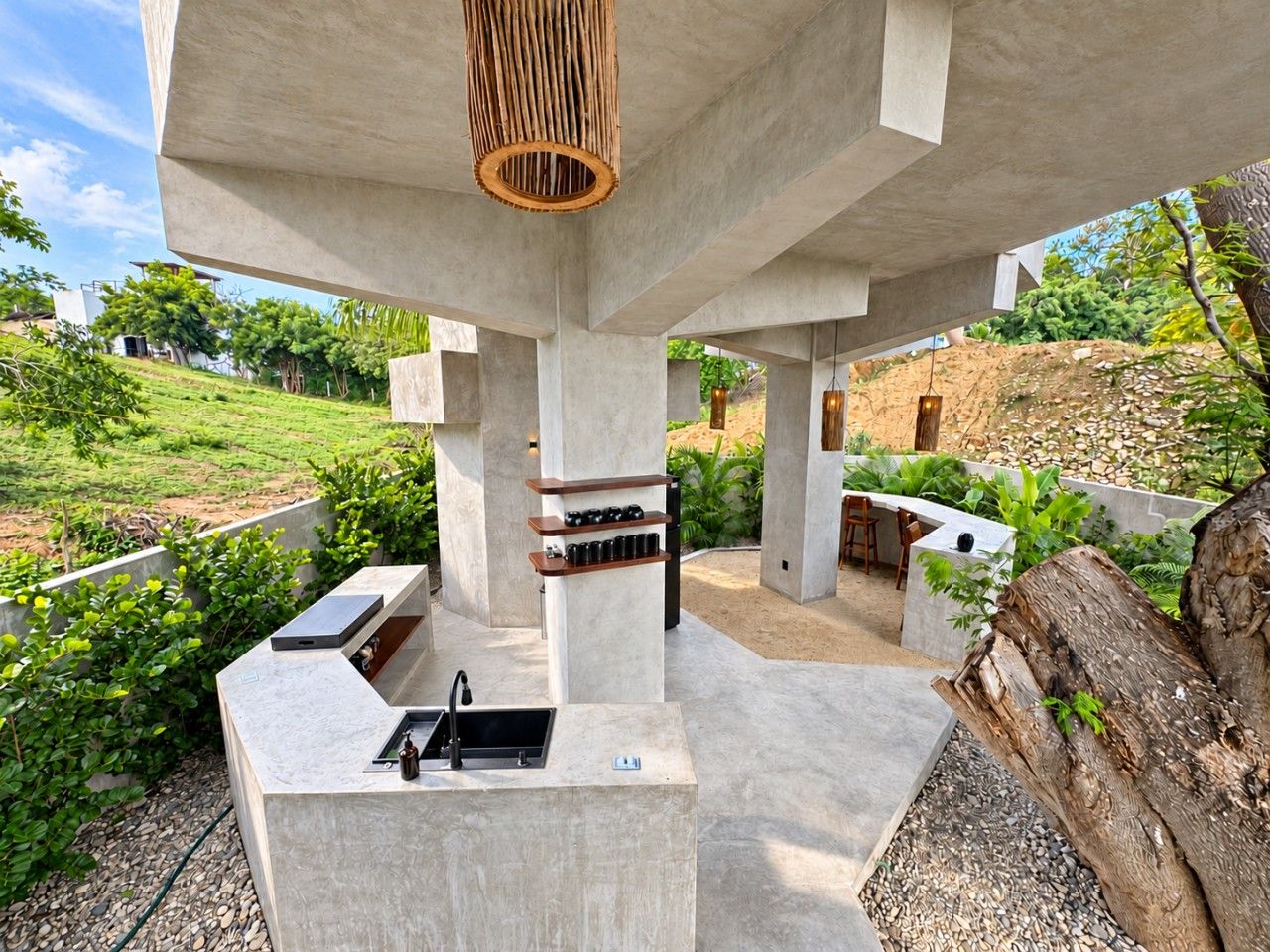An outdoor kitchen area with a built-in sink and bar seating, surrounded by greenery and a concrete structure.
