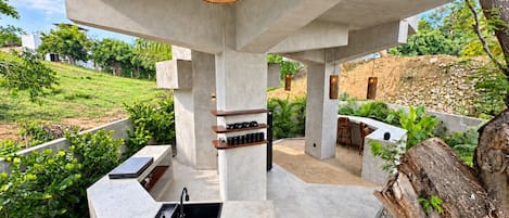 An outdoor kitchen area with a built-in sink and bar seating, surrounded by greenery and a concrete structure.