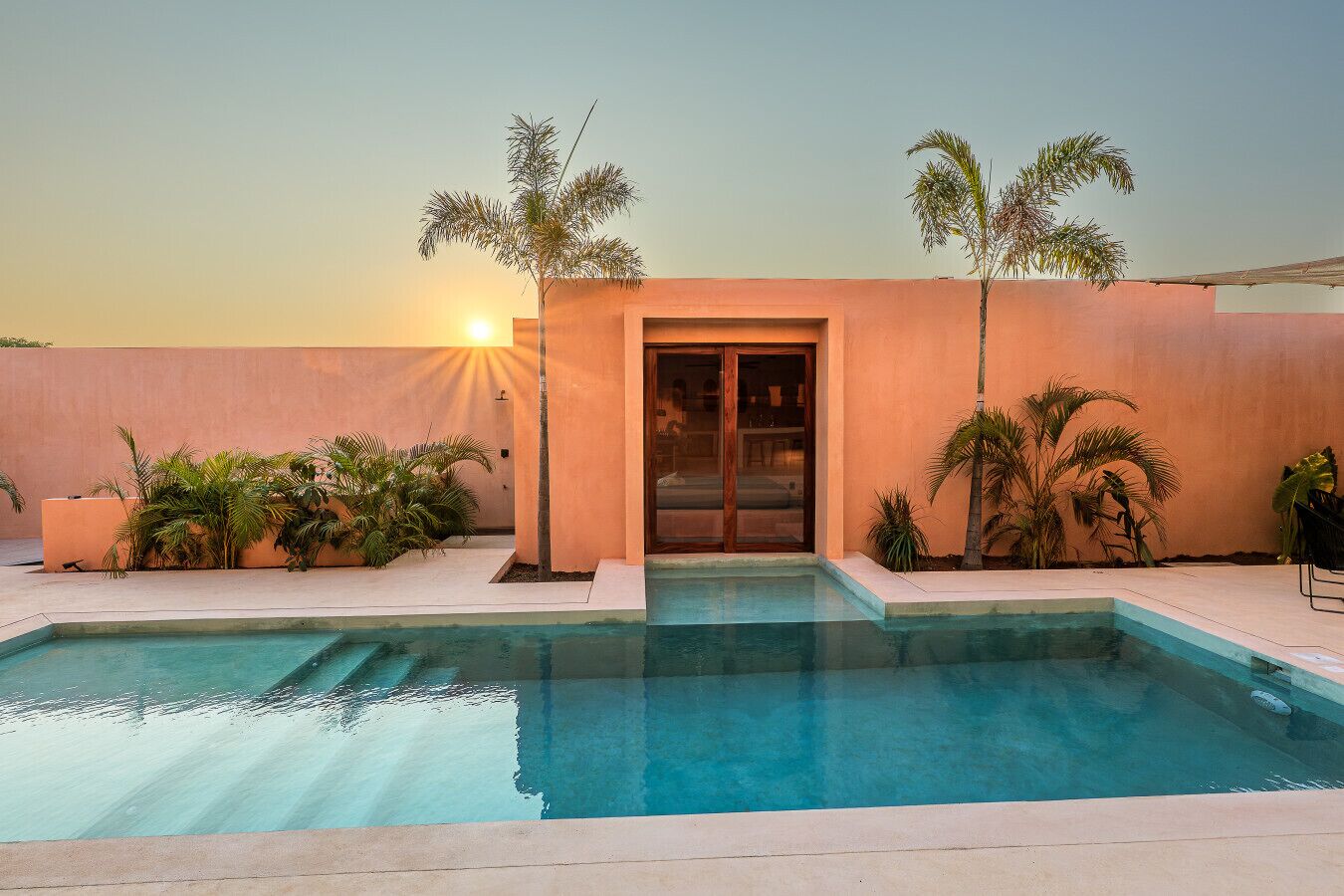 Une piscine avec un coucher de soleil en arrière-plan, des palmiers et un bâtiment doté d’une porte coulissante.